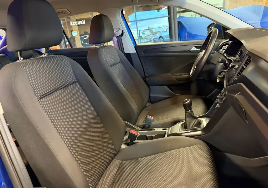 Vue intérieure côté conducteur du Volkswagen T-Roc bleu, montrant sièges tissu, volant cuir et levier de vitesse manuel.