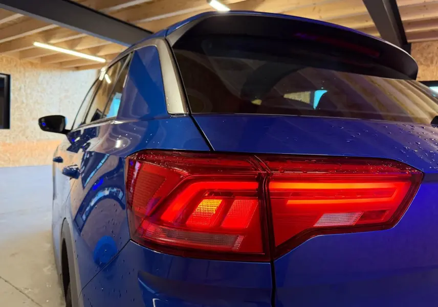 Vue 3/4 arrière gauche du Volkswagen T-Roc bleu avec feux arrière LED allumés dans un garage.