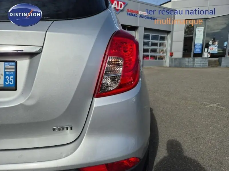 Vue arrière droite d'un Opel Mokka X gris minéral avec feux arrière et logo CDTI visibles, devant un garage multimarque.