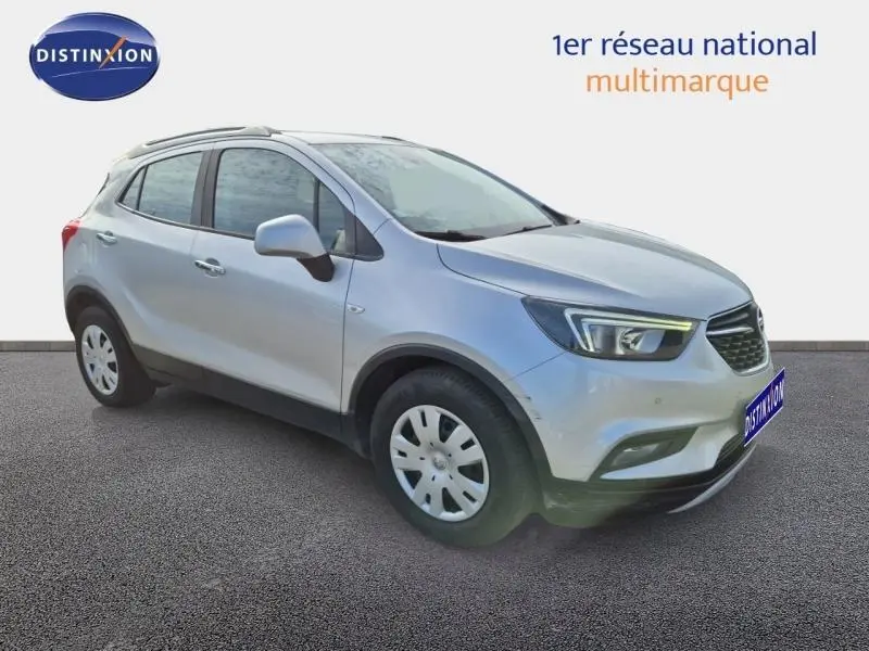 Opel Mokka X gris minéral vue 3/4 avant droit, avec jantes acier et barres de toit visibles.