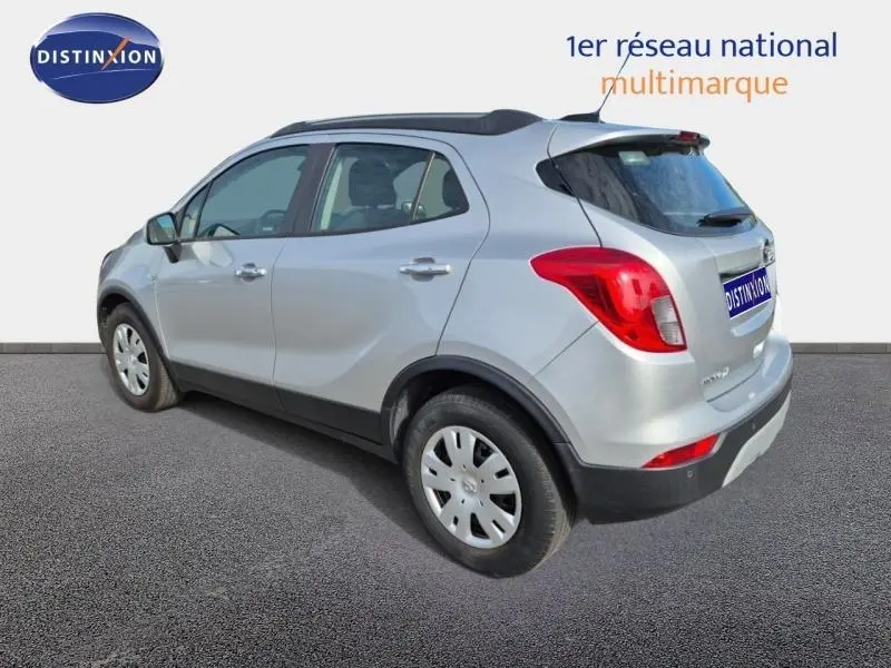 Opel Mokka X gris minéral vue 3/4 arrière droit, avec barres de toit et jantes acier.