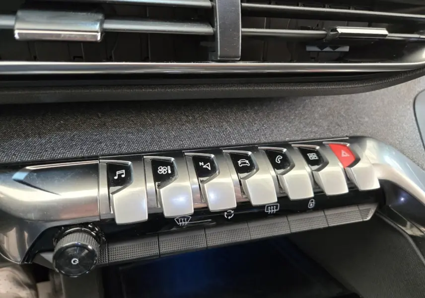 Gros plan sur la rangée de boutons argentés et noirs de la console centrale du Peugeot 5008 gris clair, avec bouton rouge d’alerte.
