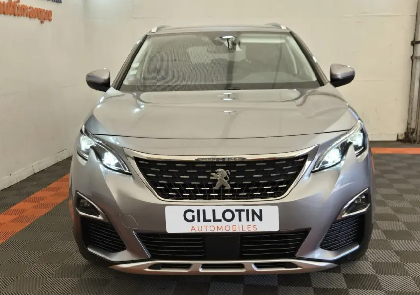Vue frontale d'un Peugeot 5008 gris clair avec phares LED allumés et calandre chromée visible en intérieur.