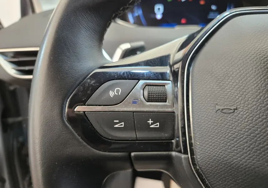 Gros plan sur les commandes gauche du volant cuir noir du Peugeot 5008 gris clair, avec boutons multifonctions et molette.