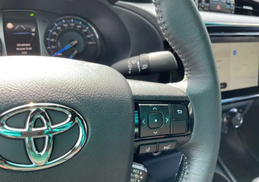 Vue rapprochée du volant cuir noir du Toyota Hilux 2025 blanc, avec commandes intégrées et tableau de bord numérique.
