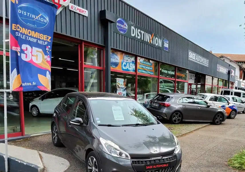 Peugeot 208 gris foncé métal vue 3/4 avant droit, garée devant un garage avec enseigne Sage Automobile.