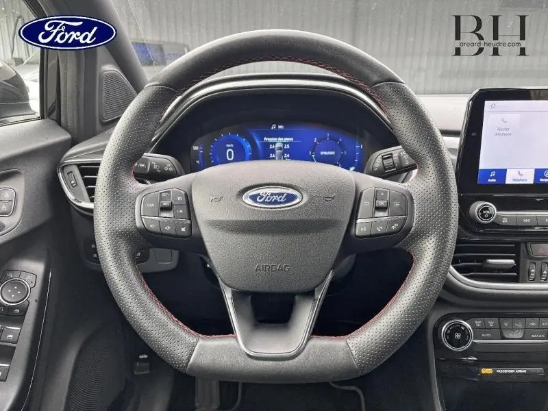 Vue centrée sur le volant sport noir avec surpiqûres rouges du Ford Puma 1.5 EcoBlue, tableau de bord digital et écran tactile à droite.