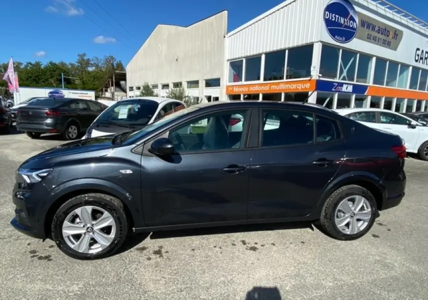 Profil côté gauche d'une Dacia Logan gris foncé 2022 garée devant un garage sous un ciel bleu.