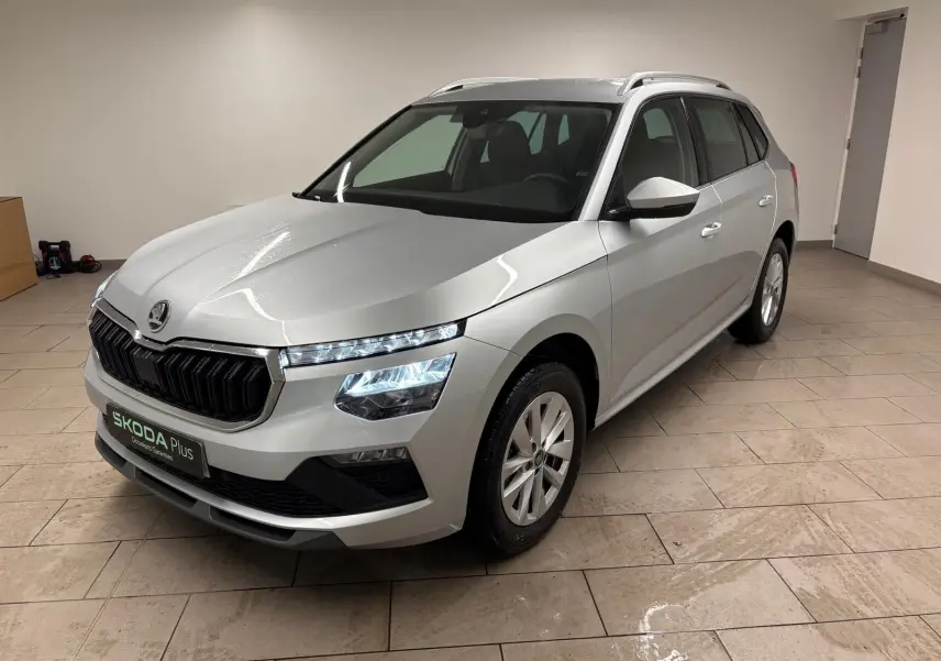 Vue 3/4 avant d'un Skoda Kamiq gris argent métallisé, avec phares allumés et calandre noire distinctive.
