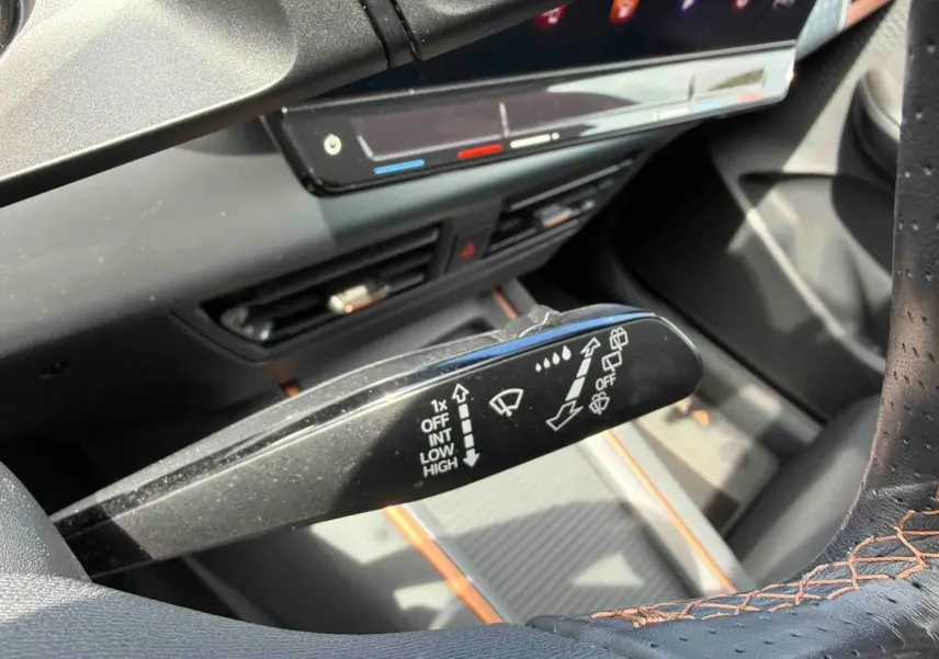 Gros plan sur la commande d’essuie-glace à gauche du volant perforé noir avec surpiqûres orange dans un CUPRA Born blanc.