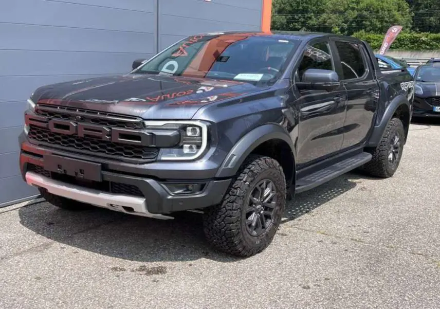 Ford Ranger Raptor 2025 gris météore en 3/4 avant droit, avec calandre imposante et pneus tout-terrain marqués.