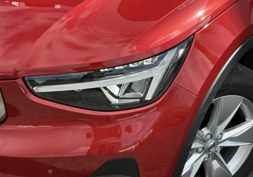 Gros plan sur le phare avant gauche et la jante alliage 18'' du Volvo XC40 rouge Fusion 2024.