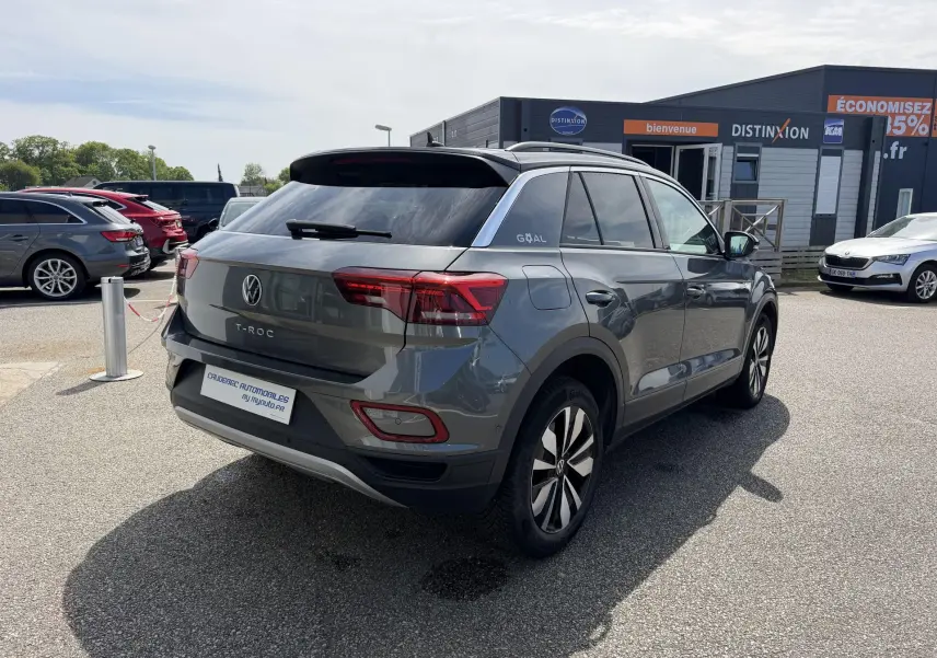 Vue 3/4 arrière droite d'un Volkswagen T-Roc gris avec barres de toit noires, stationné en extérieur devant un bâtiment commercial.
