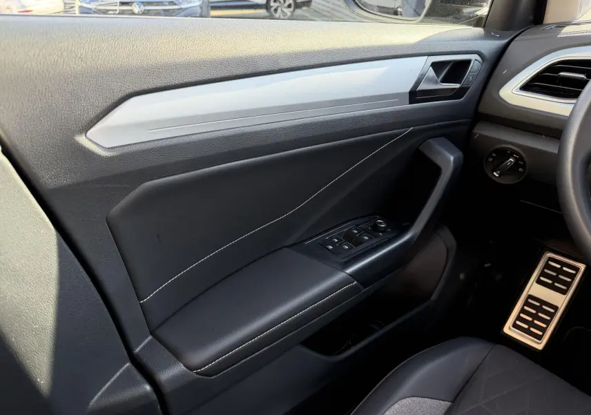 Vue intérieure côté conducteur du Volkswagen T-Roc 2025, montrant la portière noire avec commandes électriques et surpiqûres blanches.
