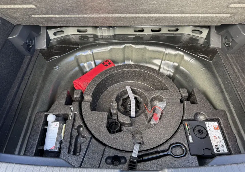 Vue plongeante sur le coffre ouvert du Volkswagen T-Roc noir, montrant le rangement sous plancher avec kit de secours et outils.
