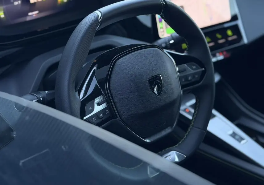Gros plan sur le volant cuir perforé noir avec logo Peugeot, tableau de bord numérique et console centrale du Peugeot 408 Hybrid 2025.