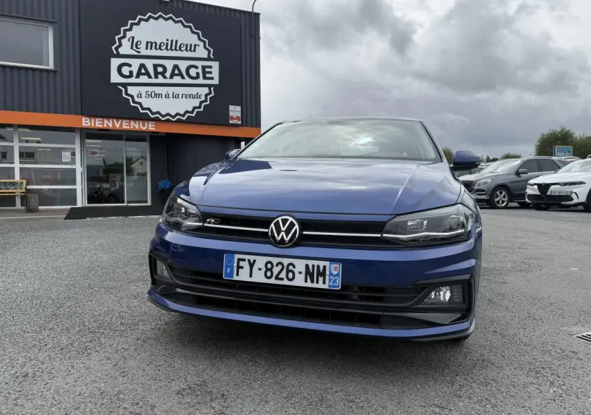Vue de face d'une Volkswagen Polo 1.0 TSI R-Line bleu métallisé avec calandre et badge R visibles, stationnée devant un garage.