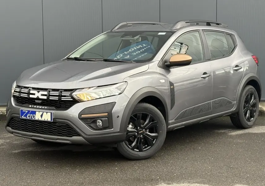 Dacia Sandero Stepway TCE 110 gris schiste en 3/4 avant droit avec jantes noires et barres de toit grises.
