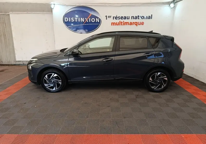 Profil côté gauche d'une Hyundai Bayon gris foncé 2023, avec jantes alliage et toit noir en arrière-plan intérieur showroom.
