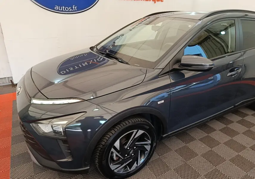Vue 3/4 avant gauche d'une Hyundai Bayon gris foncé avec jantes alliage et badge hybride visible sur l'aile avant.