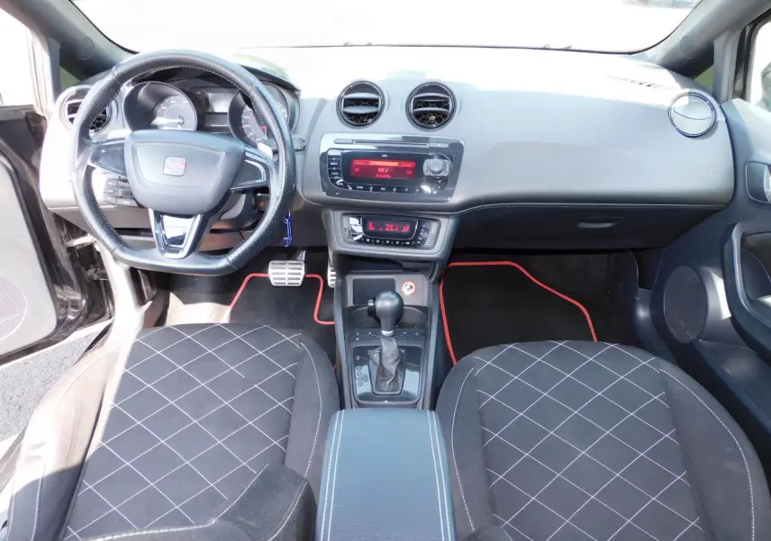 Intérieur avant de la SEAT Ibiza Cupra noire de 2011, volant cuir, console centrale avec levier de vitesse automatique et tapis noirs à liseré rouge.