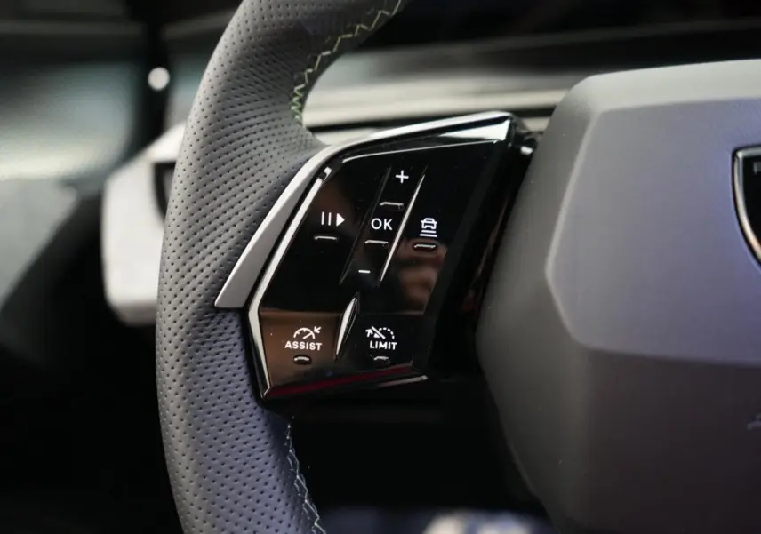 Gros plan sur les commandes gauche du volant perforé noir du Peugeot 3008 hybride 2026, avec boutons d'assistance et limiteur.