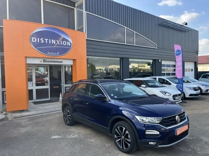 Volkswagen T-Roc bleu foncé en 3/4 avant droit, garé devant un concessionnaire avec plaque "OCCASION" visible.