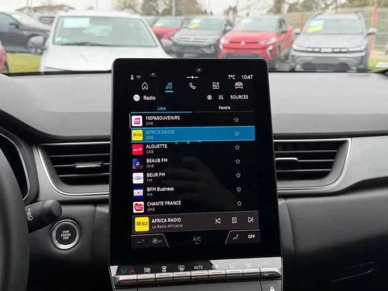 Vue intérieure centrée sur l'écran tactile du tableau de bord du Renault Captur 2025, avec vue floue de plusieurs Captur rouges à l'extérieur.