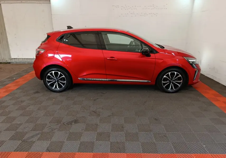 Profil droit d'une Renault Clio rouge 2024 avec jantes alliage et feux LED distinctifs, sur sol damier gris et orange.