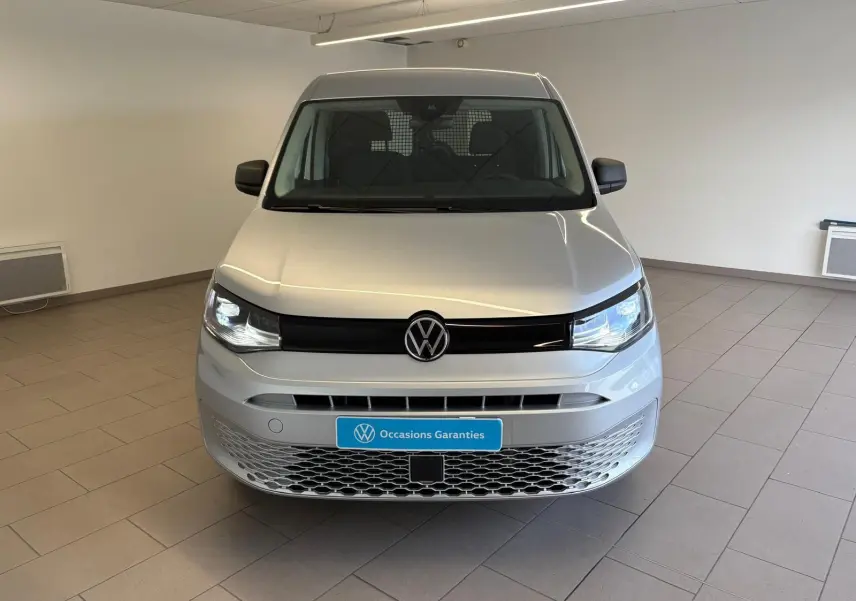 Vue frontale d'un Volkswagen Caddy 2025 gris argent avec calandre noire et plaque "Occasions Garanties" en intérieur.