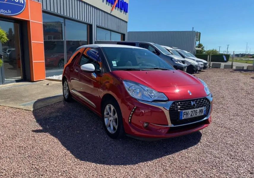 DS3 rouge avec toit blanc, vue 3/4 avant droit, garée devant un bâtiment commercial sous un ciel bleu.