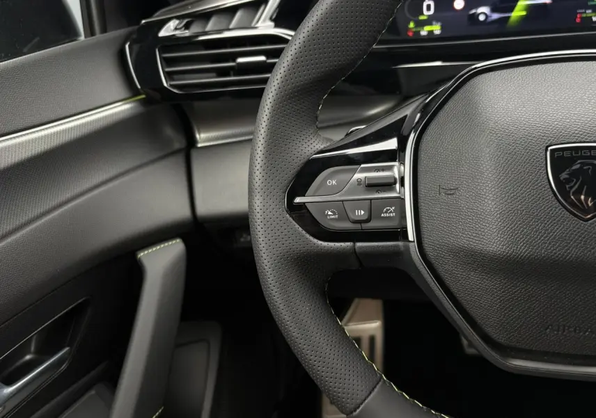 Gros plan sur le volant perforé noir avec surpiqûres vert Adamite et partie gauche du tableau de bord gris et noir du Peugeot 308 SW Hybrid.