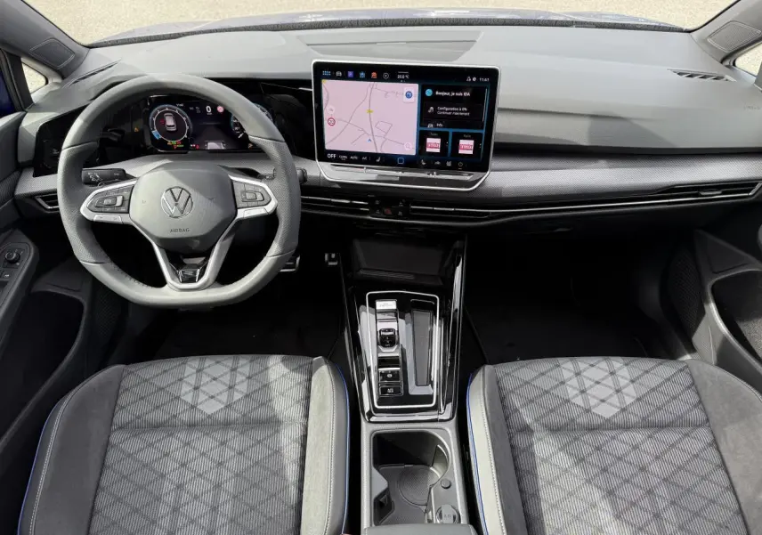 Vue intérieure avant de la Volkswagen Golf VIII R-Line 2025, tableau de bord noir avec écran tactile central et sièges tissu gris à motifs.