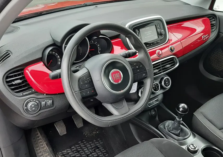 Vue intérieure côté conducteur d'une FIAT 500X rouge, sièges gris avec logo 500 brodé et tableau de bord assorti rouge.