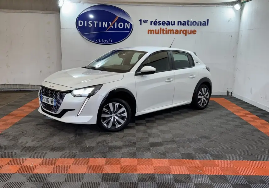 Peugeot 208 électrique blanche vue 3/4 avant droit dans un garage avec sol noir et orange.