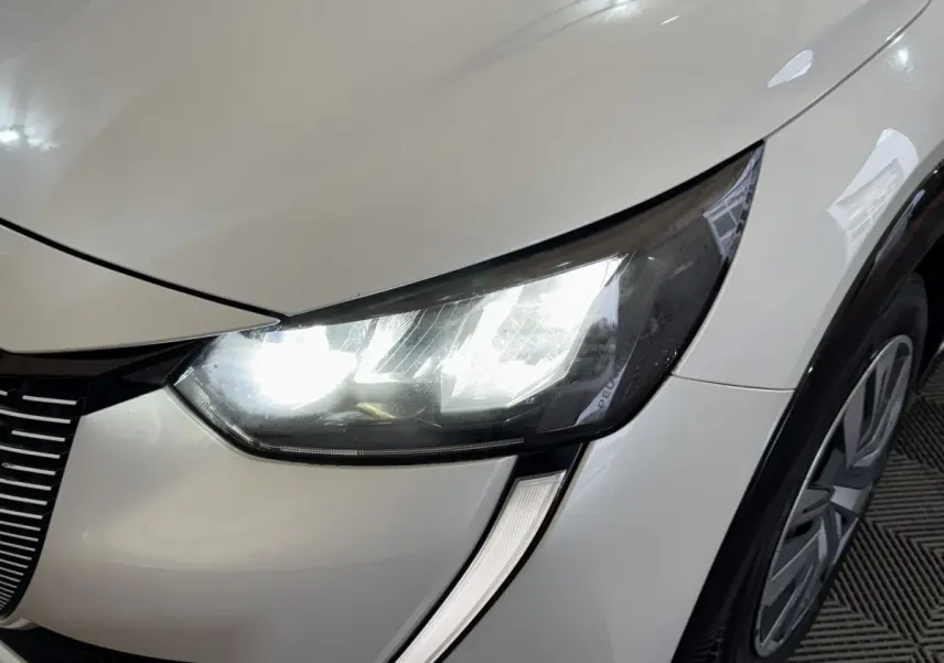 Gros plan sur le phare avant droit allumé d'une Peugeot 208 électrique blanche, avec partie de la roue visible.
