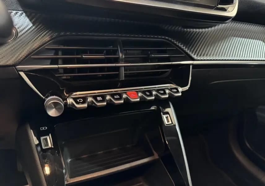 Gros plan sur la console centrale noire de la Peugeot 208 électrique, avec boutons métalliques et finition carbone.