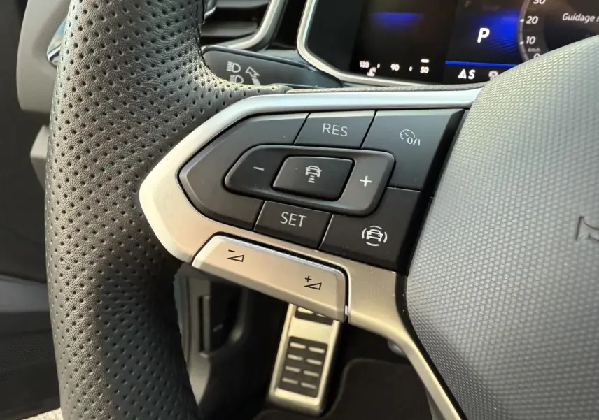 Gros plan sur les commandes multifonctions à gauche du volant perforé d’un Volkswagen T-Roc 2025, avec pédale en acier visible.