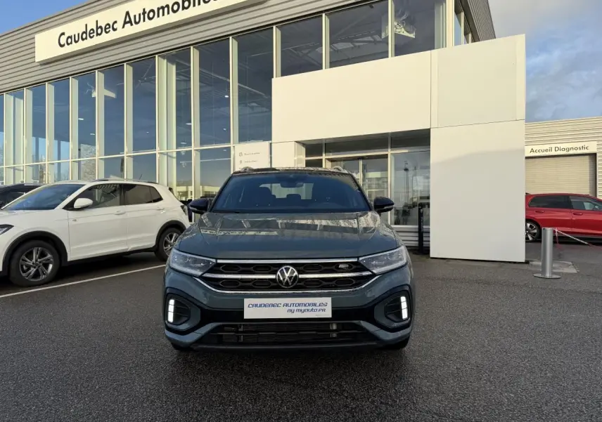 Vue avant d'un Volkswagen T-Roc bleu garé devant un concessionnaire avec calandre et feux LED visibles.