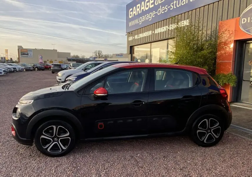 Profil côté gauche d'une Citroën C3 noire avec toit et rétroviseurs rouges, garée devant un garage.