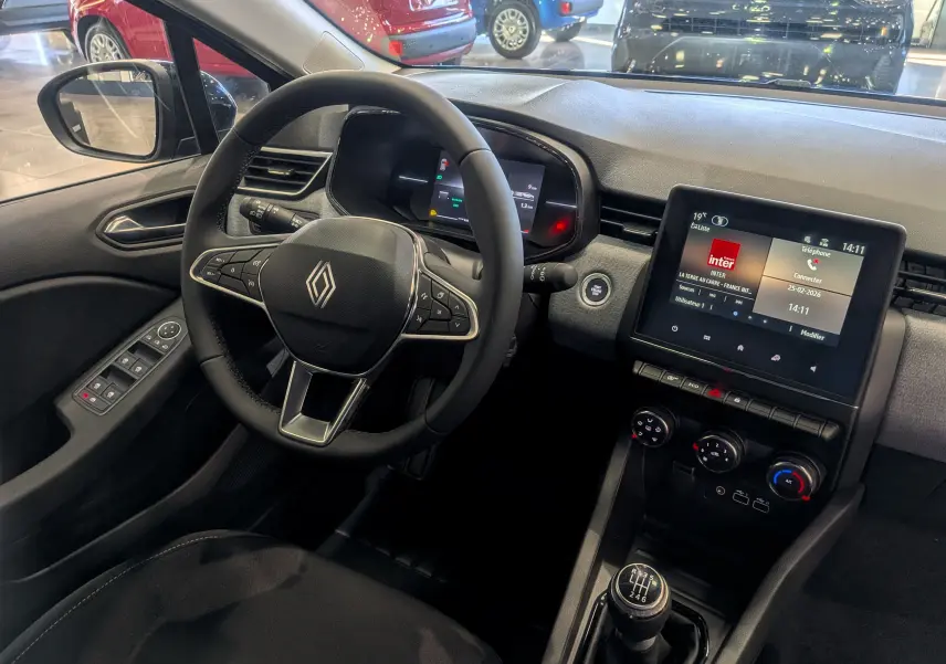 Vue intérieure côté conducteur de la Renault Clio V 2025, tableau de bord moderne avec écran tactile et volant multifonction noir.
