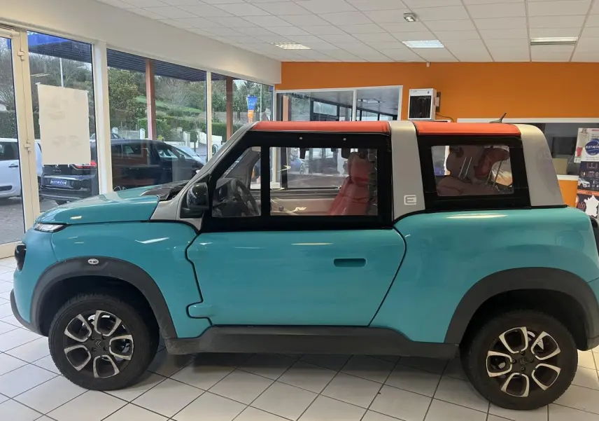 Vue de profil côté gauche d'une Citroën E-MEHARI cabriolet bleu clair avec toit en toile rouge en intérieur showroom.
