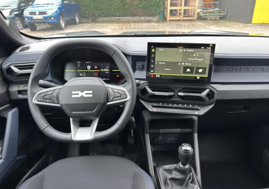 Vue intérieure du tableau de bord du Dacia Duster 2025 avec volant multifonction, écran tactile et levier de vitesses manuel.