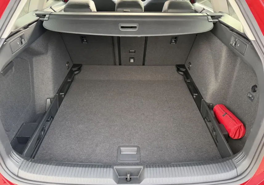 Coffre ouvert vu de l'arrière d'une Volkswagen Golf SW rouge, avec tapis gris et trousse de secours rouge à droite.