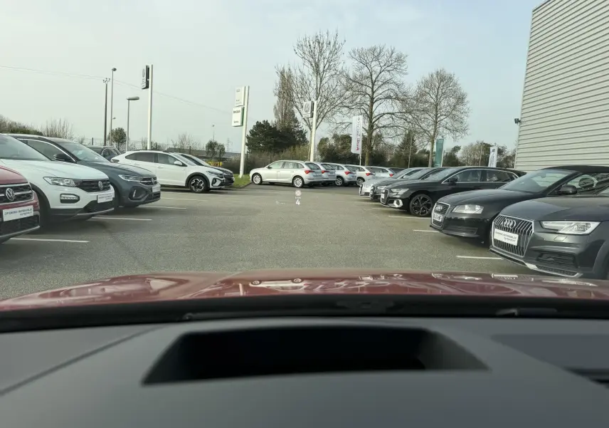 Vue depuis l'habitacle d'une Volkswagen Golf SW rouge, avec le capot visible au premier plan, dans un parking extérieur.