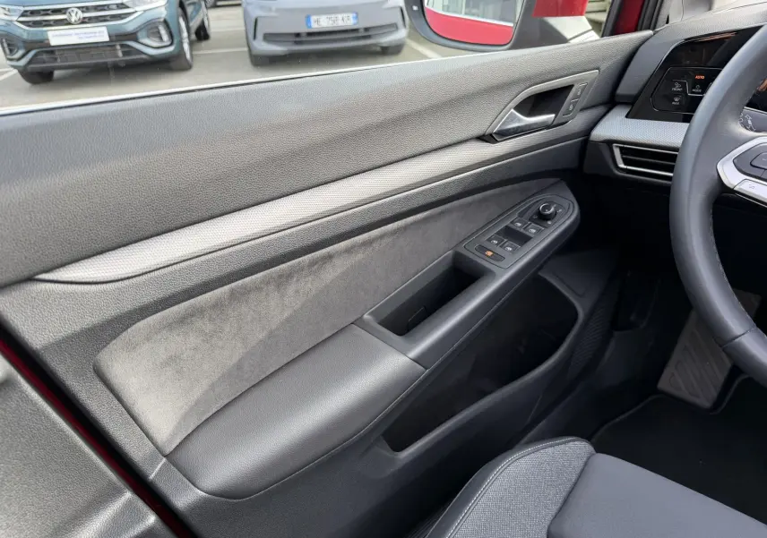 Vue intérieure côté conducteur de la porte d’une Volkswagen Golf SW rouge, avec commandes électriques des vitres et garniture grise.