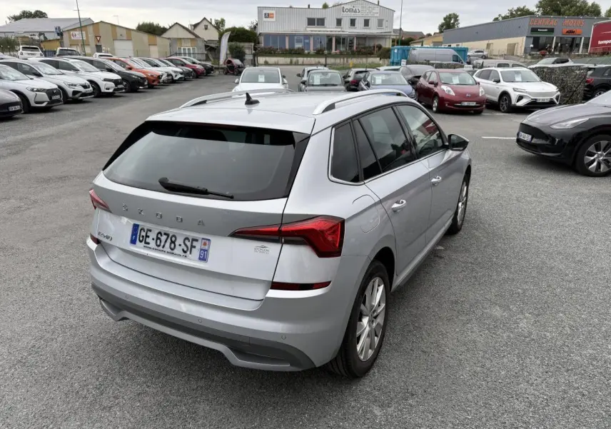 Vue 3/4 arrière droite d'un Skoda Kamiq gris métallisé avec barres de toit et feux arrière LED dans un parking.