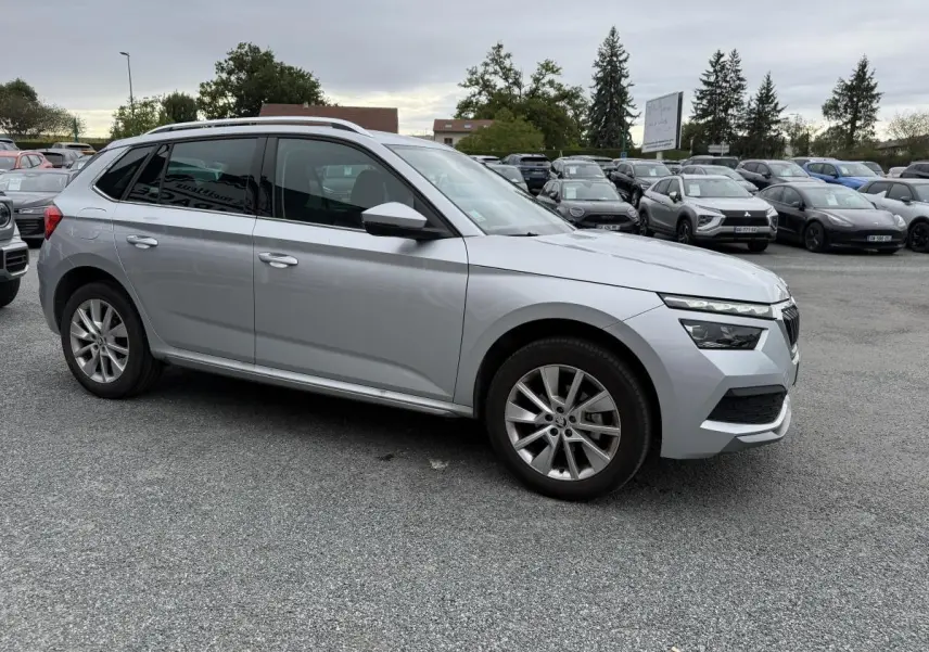 Vue profil droit d'un Skoda Kamiq gris métallisé 2022, avec jantes alliage et barres de toit argentées sur parking.