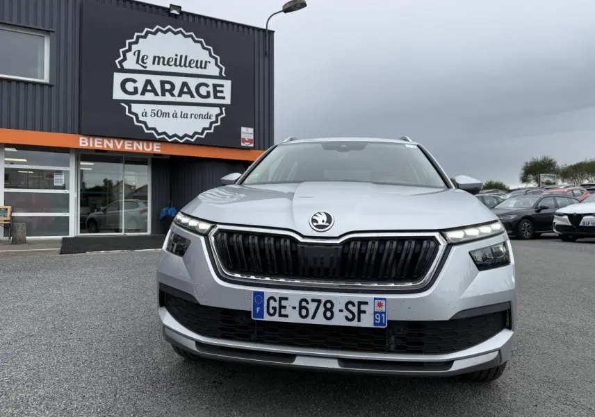Vue de face d'un Skoda Kamiq gris métallisé avec calandre noire et phares LED allumés devant un garage.