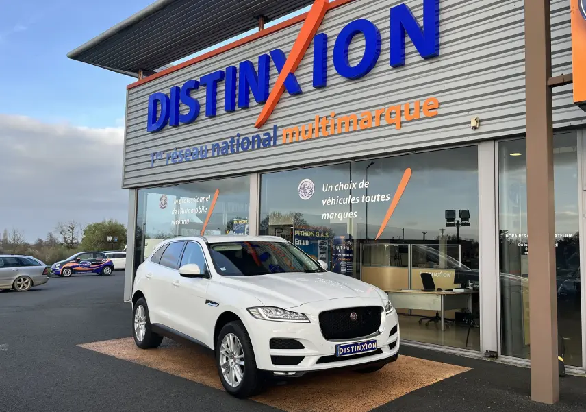 Jaguar F-PACE blanc en 3/4 avant droit, stationné devant un showroom Distinxion avec une calandre noire distinctive.